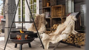 Una chimenea con soporte metálico, una terraza de madera y un interior rústico de cabaña con estantes de madera y un aparador de vidrio.