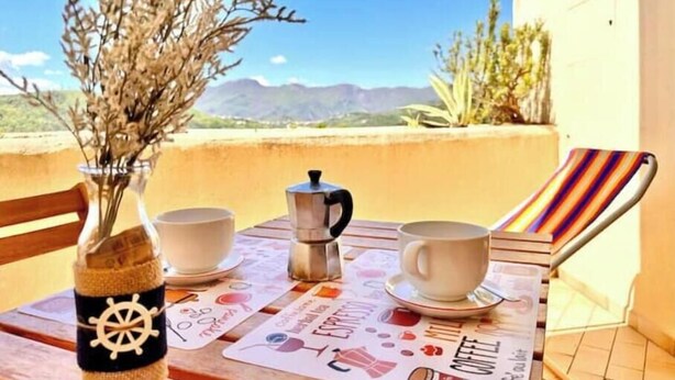Une table avec une cafetière, deux tasses, une chaise à rayures et une vue sur les montagnes.