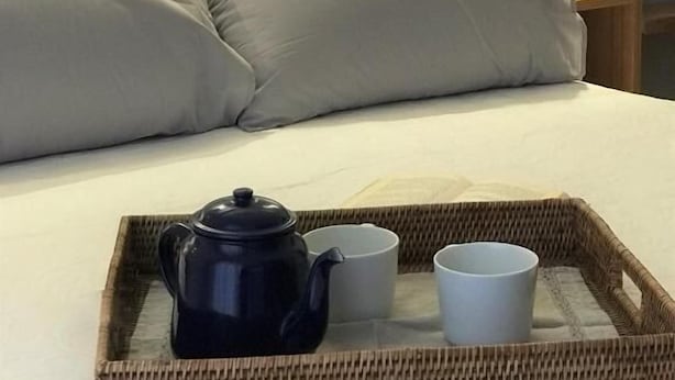 A neatly made bed with pillows, a tray with a teapot and cups, and a bedside table with a lamp.