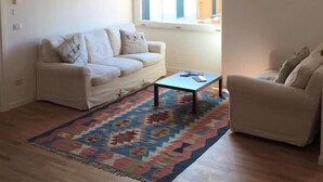 A living room with a sofa, a coffee table, a patterned rug, a hanging light fixture, and a window with a view of buildings.