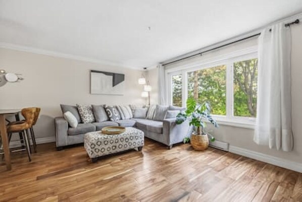 Un salon avec un canapé gris, un pouf à motifs, un plancher en bois, un coin repas avec une chaise en bois et une grande fenêtre avec des rideaux.