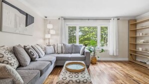 Ein Wohnzimmer mit grauem Sofa, einem gemusterten Hocker, einem Beistelltisch, einer Stehlampe und einem Bücherregal.