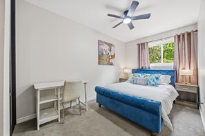 Une chambre à coucher comprenant un lit, un bureau, une chaise, un ventilateur de plafond, des œuvres d’art, une fenêtre avec des rideaux et des lampes de chevet.