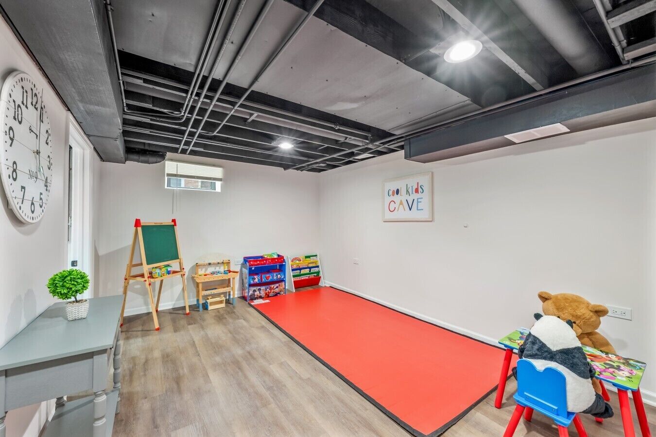 A room with a red carpet, a green easel, colorful storage units, a clock, and a teddy bear on a chair.