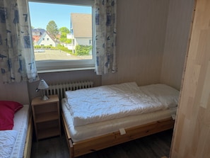A bedroom with a single bed, a nightstand, a lamp, a window with curtains, and a radiator.