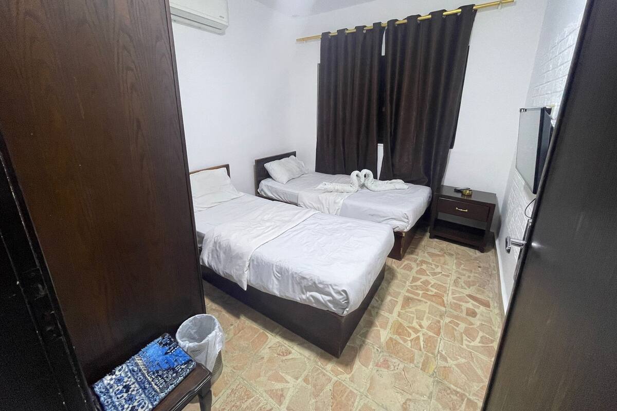 A hotel room with two beds, a small table, a trash bin, and a window with curtains.