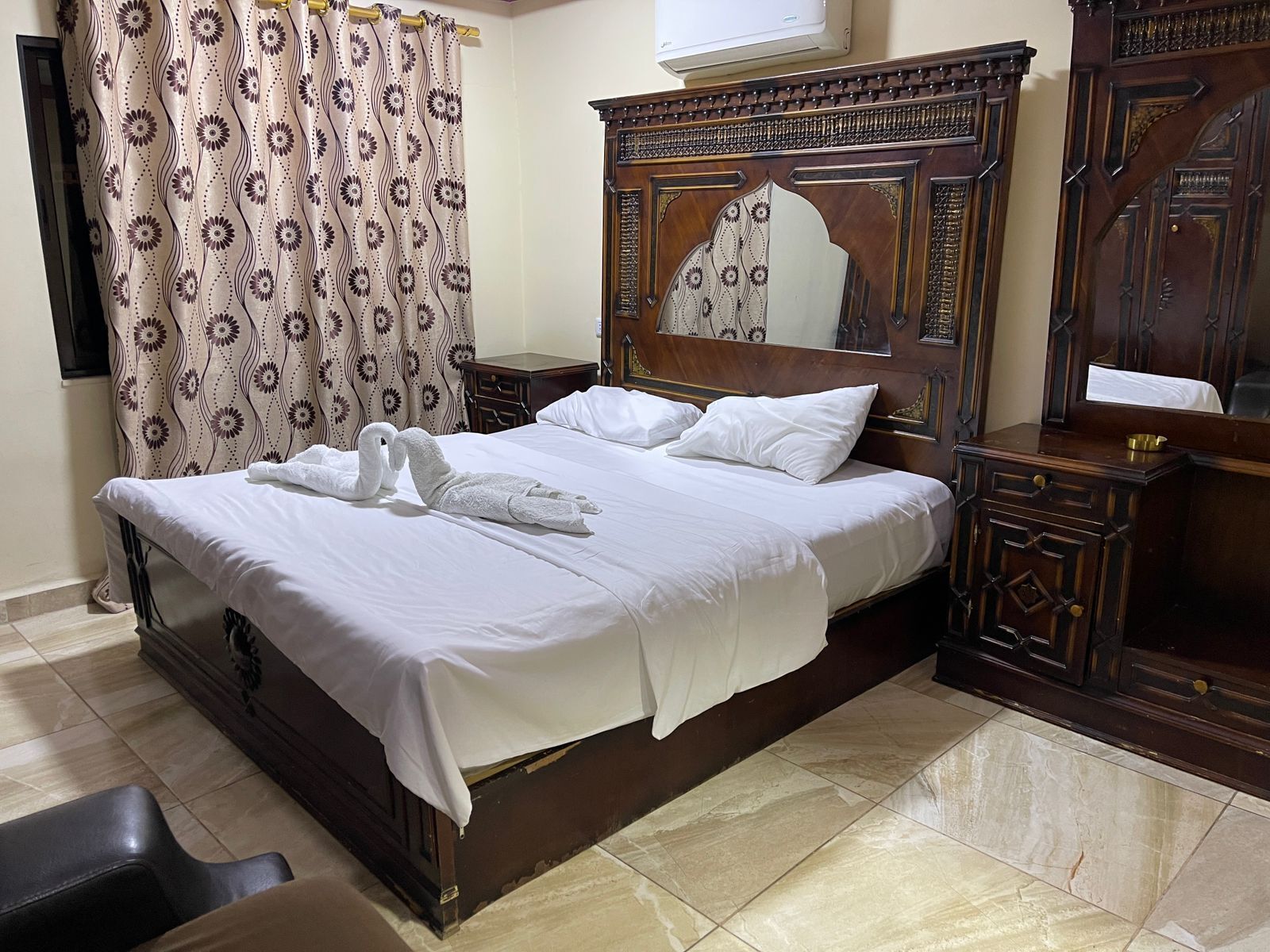 A neatly made bed with white linens and two swan-shaped towels, a wooden headboard with intricate carvings, a mirror, and a wooden nightstand with a brass bowl.