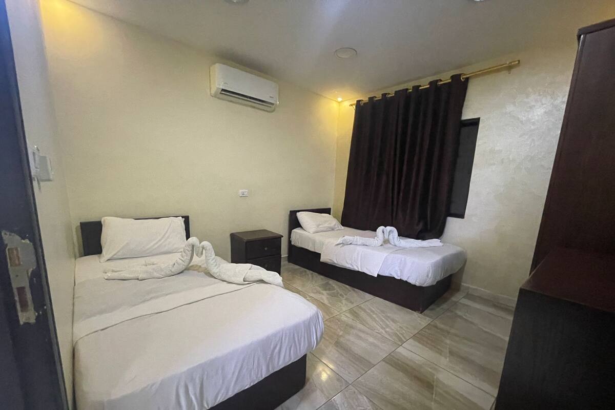 A hotel room with two beds, a nightstand, an air conditioning unit, and a window with curtains.