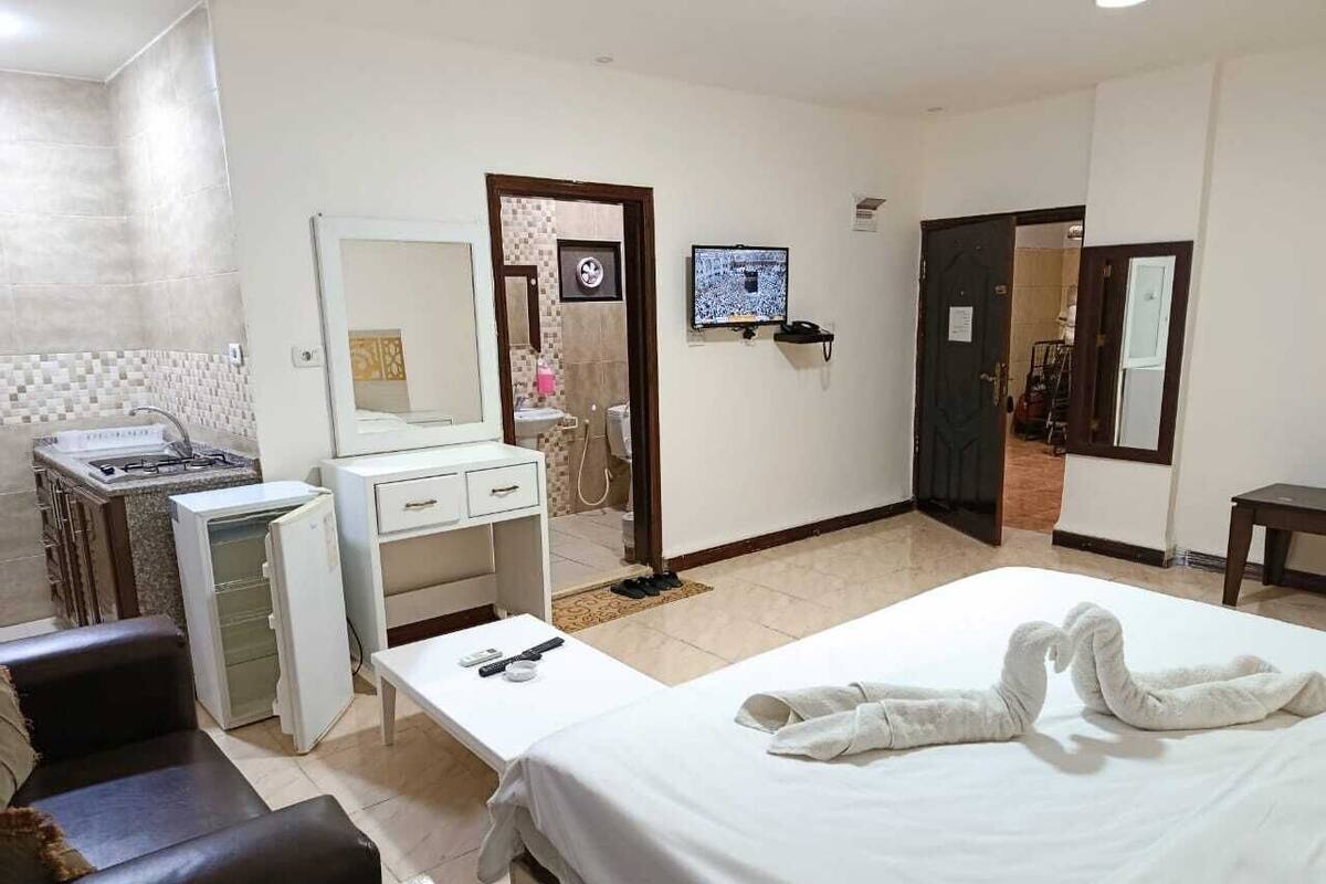 A hotel room with a bed, a desk with a chair, a small refrigerator, a mirror, a TV, and a bathroom visible through an open door.