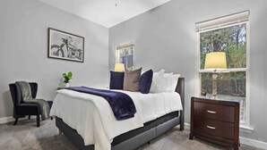 Un dormitorio con cama, una silla, una mesita de noche, una ventana y un ventilador de techo.