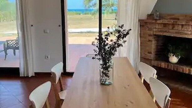Een eethoek met een houten tafel, witte stoelen, een vaas met bloemen, een open haard en uitzicht op het strand door grote schuifdeuren.