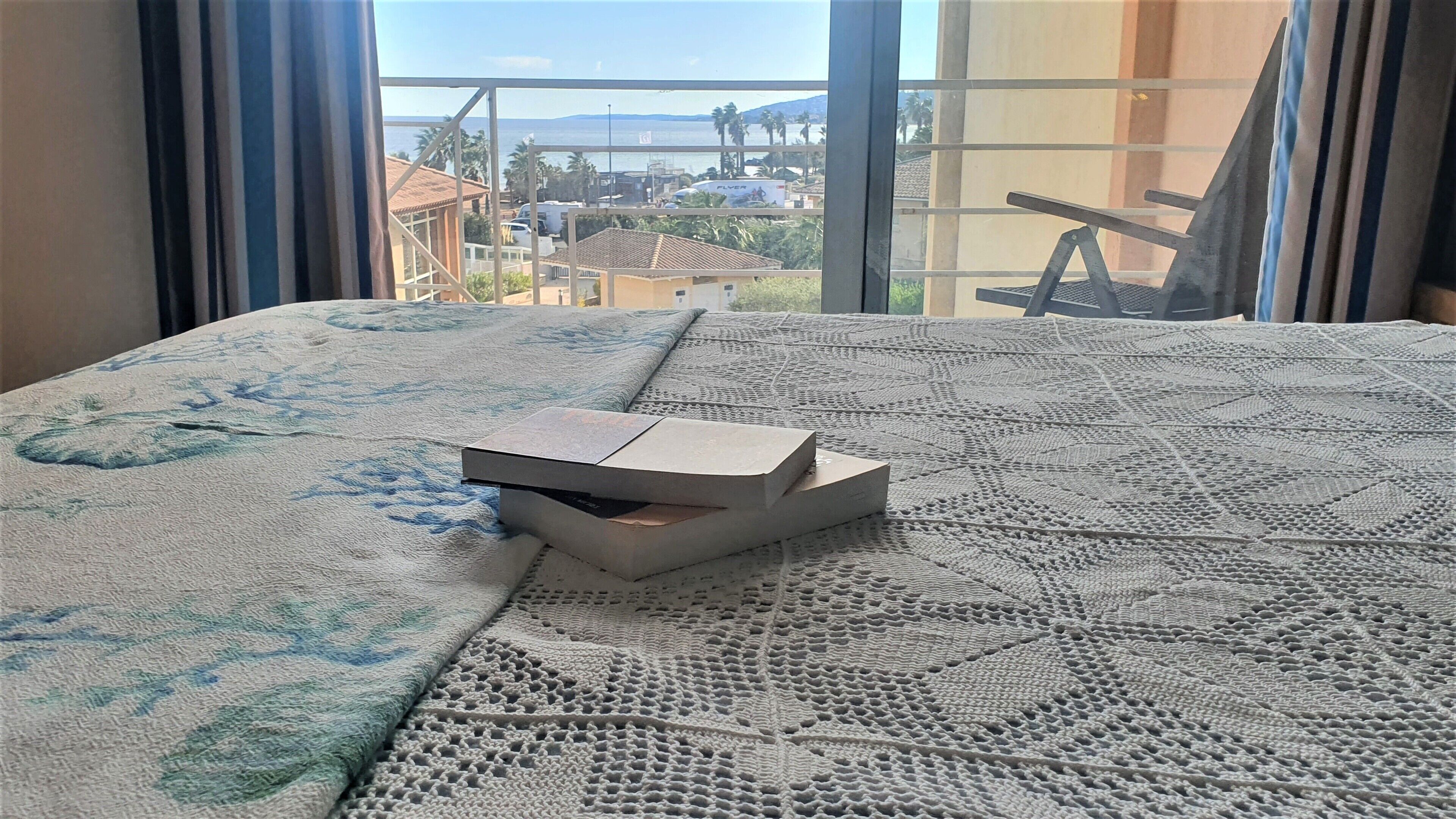 Un letto con una coperta a fantasia, un oggetto su di esso e vista di edifici e palme attraverso una finestra.