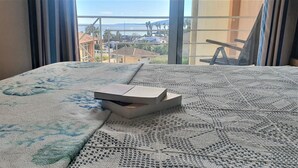 Una cama con una colcha estampada, una caja sobre la cama y vistas de edificios y palmeras a través de una ventana.