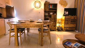 Un comedor con mesa y sillas de madera, una cocina al fondo, una estantería y un televisor.