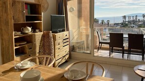 Comedor con una mesa lista para dos, un televisor sobre un mueble de madera y un balcón con vista a la playa.