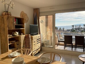 A dining area with a table set for two, a television on a wooden cabinet, and a balcony with a view of the beach. - Exceptional, 2-bedroom apartment, waterfront, sea view (Fréjus)