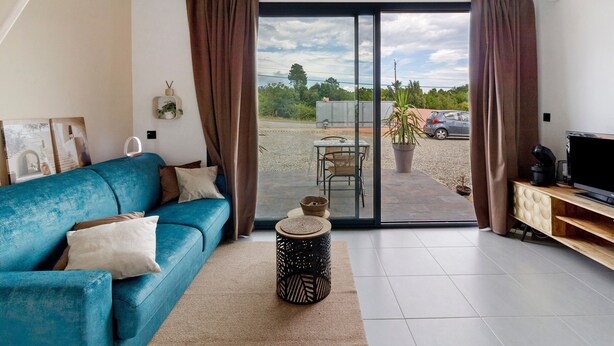A living room with a blue sofa, a small round table, a TV stand, and a sliding glass door leading to an outdoor area with a car and greenery. - Bed and Breakfast 'Vill'amore' with Sea View, Wi-Fi and Air Conditioning (Solaro)