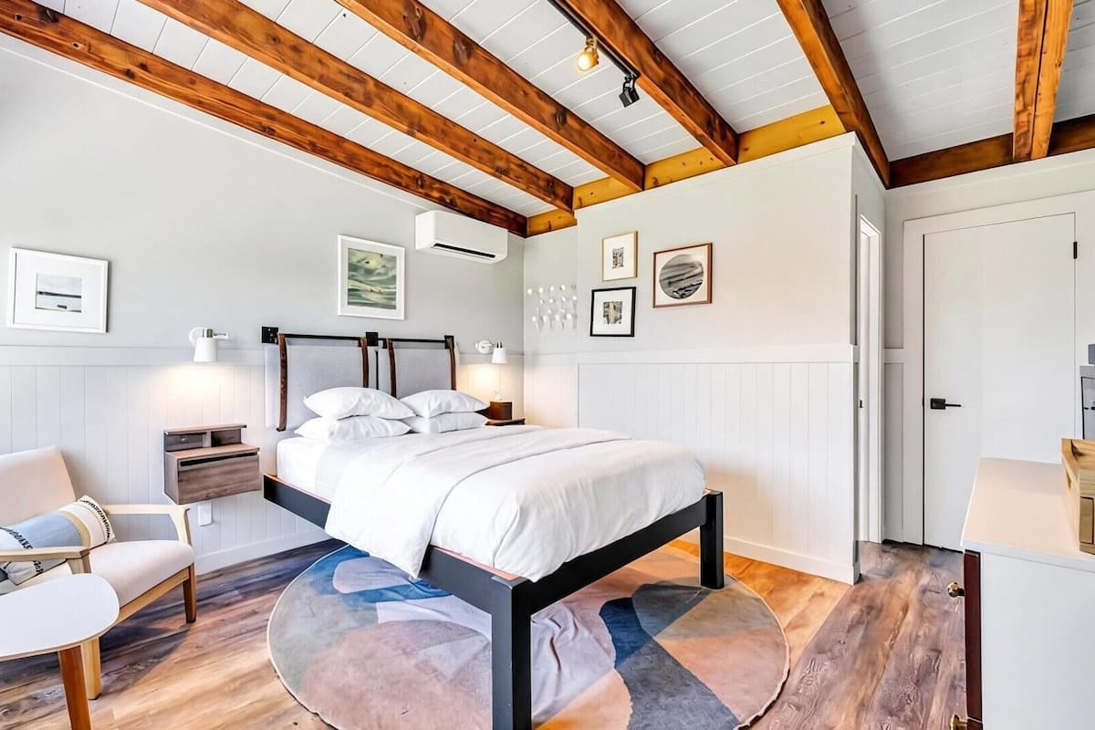Une chambre à coucher comprenant un lit, une chaise, un tapis rond, des poutres en bois et des œuvres d’art accrochées au mur.