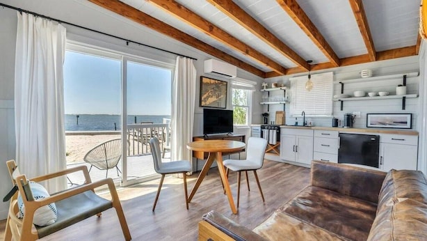 Ett rum vid stranden med en stor skjutdörr i glas, en matplats med ett runt bord, en soffa och ett kök med vita skåp och en tv.