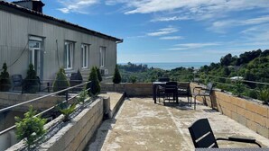 Una terraza en la azotea con vistas al mar, un edificio con techo de metal corrugado y mesas al aire libre.