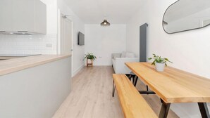 Una cocina moderna con armarios blancos, encimera de madera, una banca, una mesa de comedor, una planta en maceta, un sofá, un televisor y una lámpara colgante.