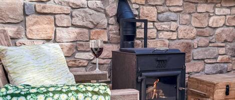 A cozy room with a stone wall, a wooden bed with a green floral blanket, a small table with a wine glass, and a black wood-burning stove.
