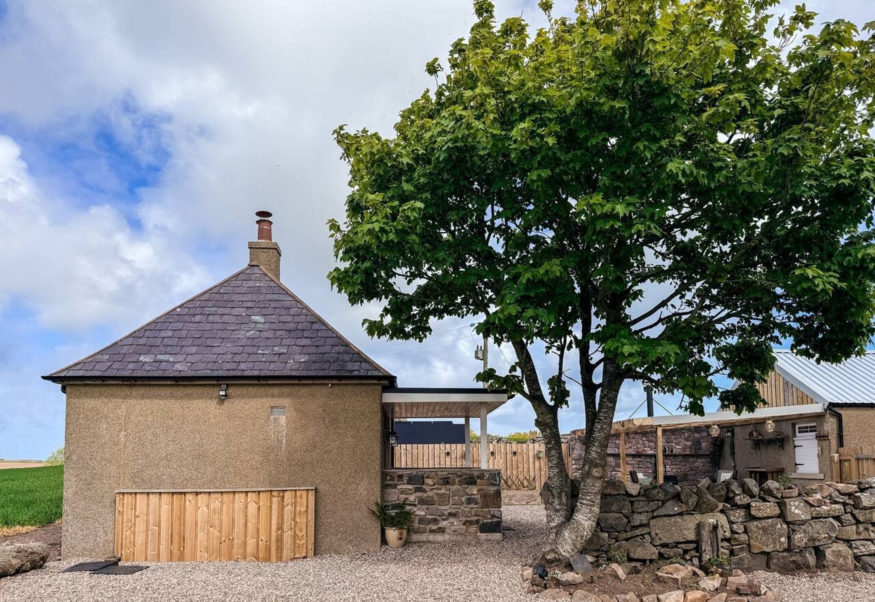 Un bâtiment rustique avec un mur en pierre, une clôture en bois et un arbre devant.