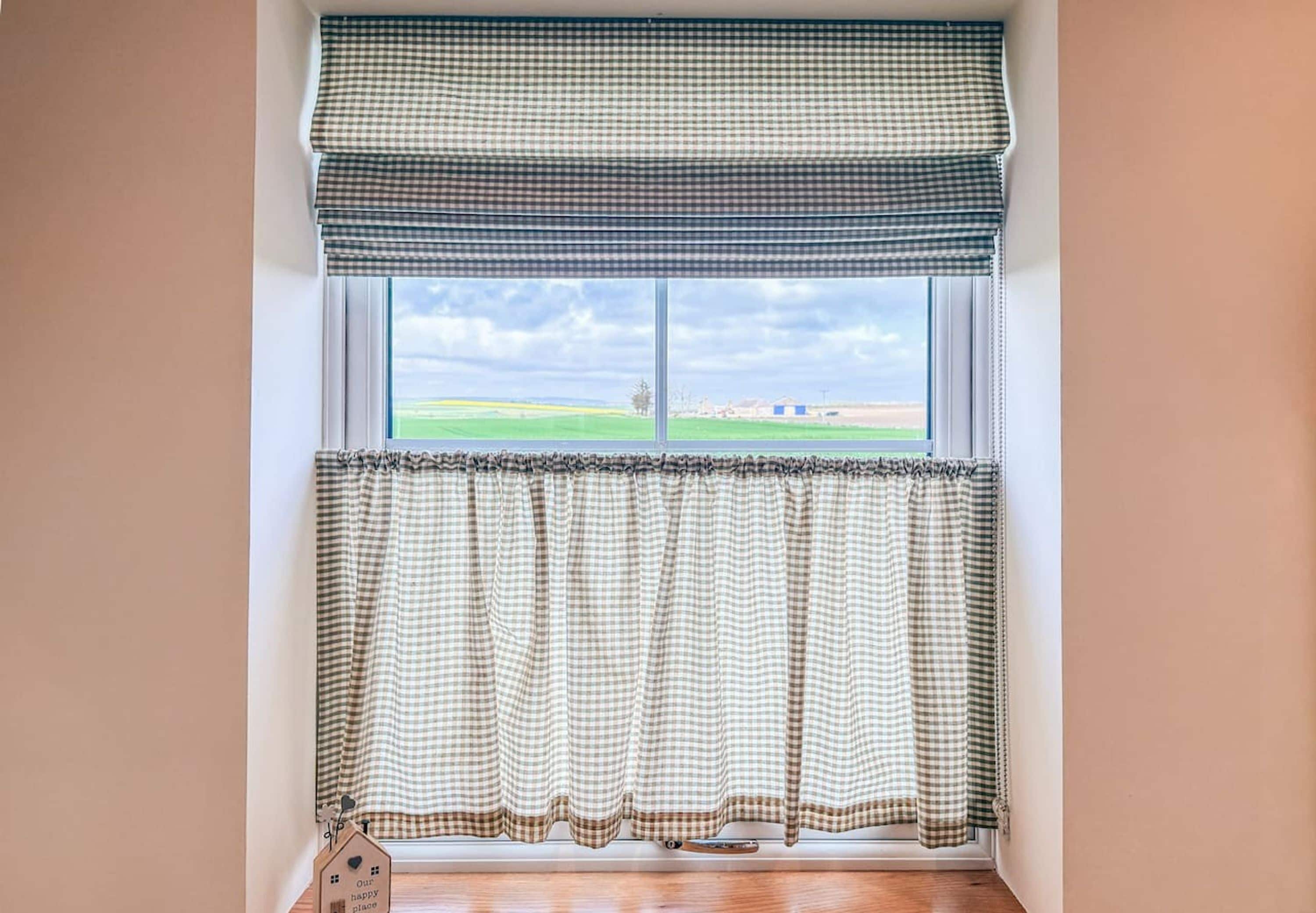 Ein Fenster mit einem karierten Vorhang und passendem Fensterteppich, Holzboden und Blick auf ein grünes Feld sowie entfernte Gebäude.
