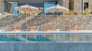 Una piscina con sillas de descanso y sombrillas, muros de piedra y un edificio moderno al fondo.