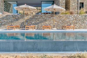 A pool with lounge chairs and umbrellas, stone walls, and a modern building in the background. - Villa Pétra - Hidden treasure (Kakapetra)