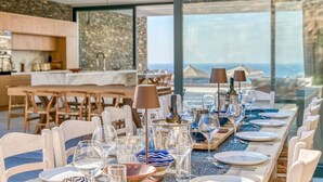 Un comedor con una larga mesa dispuesta para una comida, amplias ventanas con vista al mar y una pared de piedra al fondo.