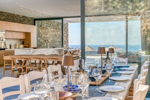 A dining area with a long table set for a meal, large windows with a view of the sea, and a stone wall in the background. - Villa Pétra - Hidden treasure (Kakapetra)