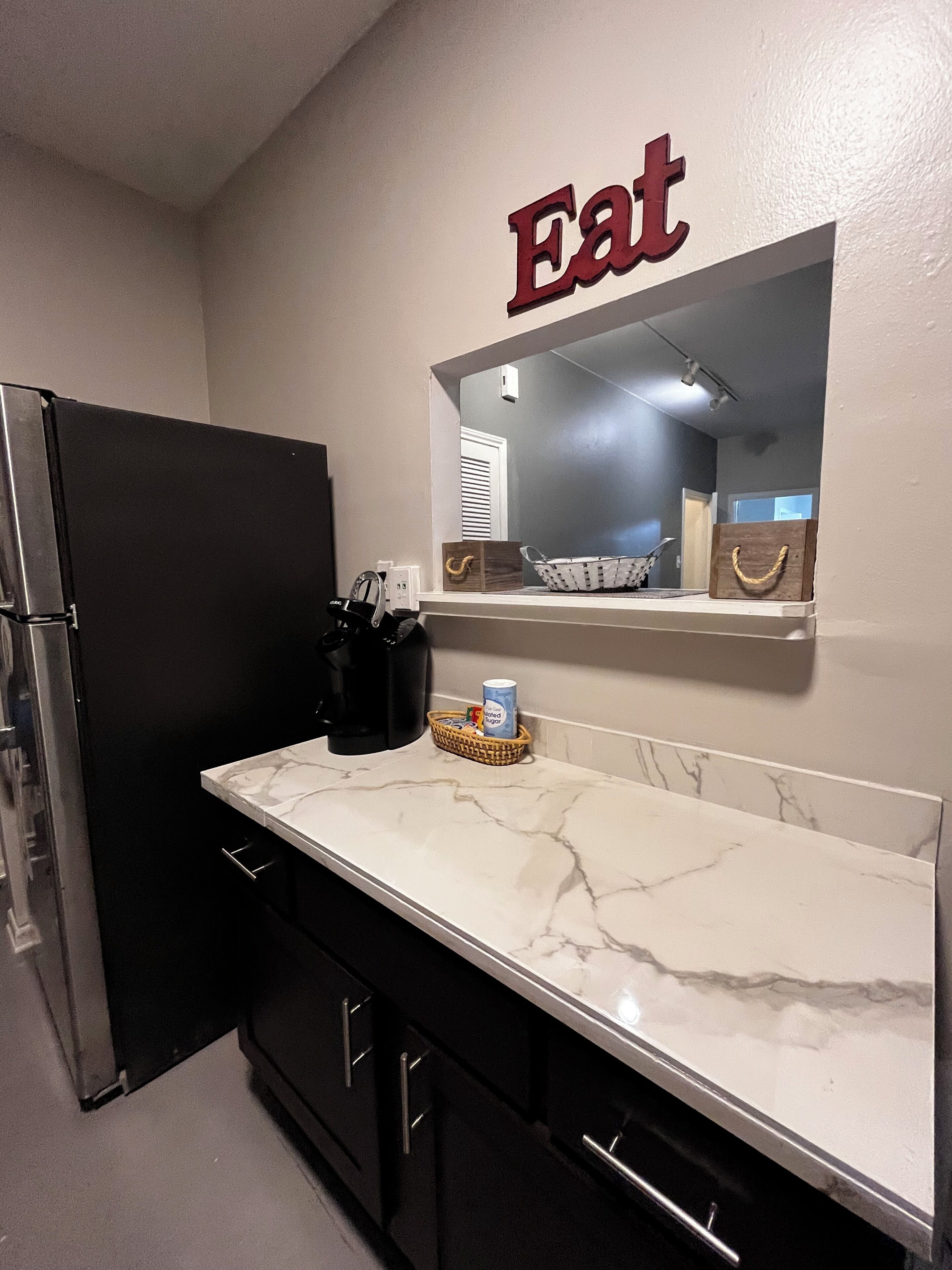 A kitchen with a refrigerator, a marble countertop, a basket with a can, and a wall-mounted mirror.