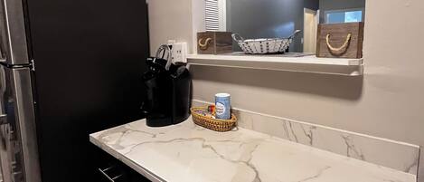 A kitchen with a refrigerator, a marble countertop, a basket with a can, and a wall-mounted mirror.