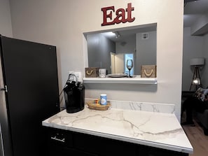 Eine Küche mit Kaffeemaschine, einer Arbeitsplatte, einem Regal mit Glas und Tasse sowie einem Schild mit der Aufschrift 'Eat'.