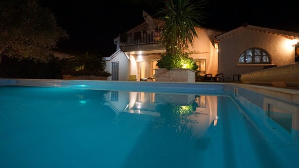 Vista noturna de uma piscina com uma casa ao fundo.