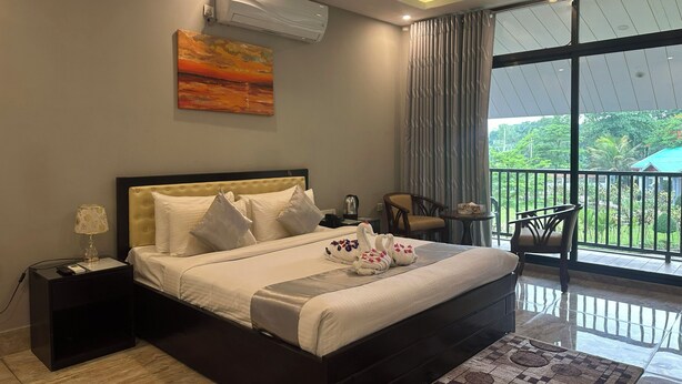 A bedroom with a bed, bedside table, lamp, air conditioning unit, artwork, and a balcony with chairs and a table. - Soaa Resort (Sirajganj)