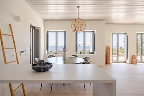 A modern dining area with a large table, chairs, a hanging light fixture, and large windows offering a view of the sea. - Amazing view of Naxos (X6W3+3G4 Paros, Greece)