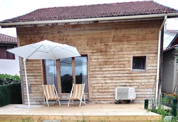 A wooden house with a tiled roof, a covered porch with two striped chairs and a white umbrella, and an air conditioning unit.