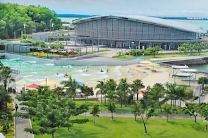 A modern building with a curved roof, a swimming pool, and a beach area with palm trees. - Coastal Magnificence Pool Workspace Parking (Darwin)