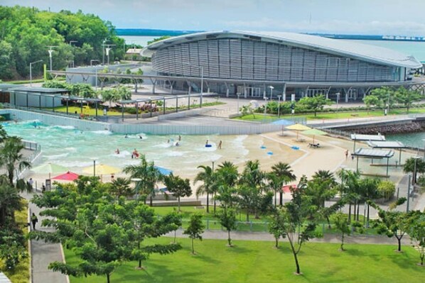 A modern building with a curved roof, a swimming pool, and a beach area with palm trees. - Coastal Magnificence Pool Workspace Parking (Darwin)