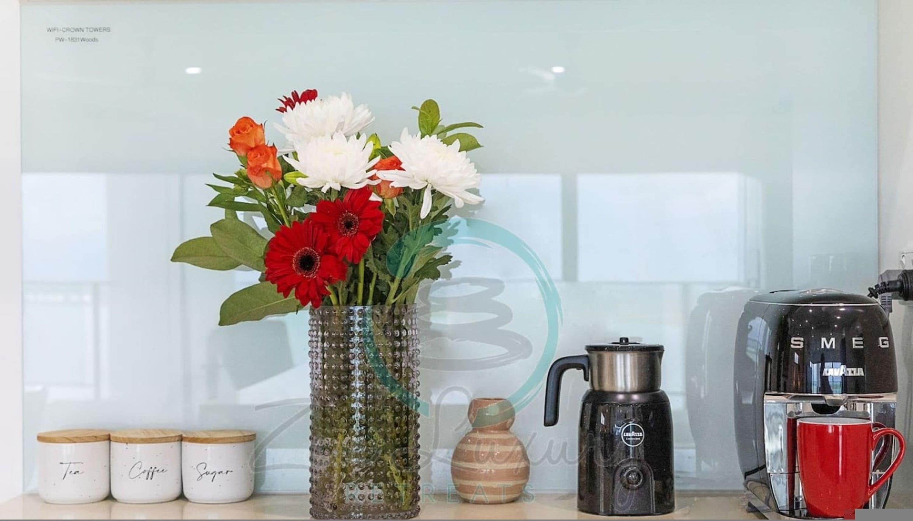 A kitchen counter with a decorative vase, a coffee maker, a red mug, and labeled containers for tea, coffee, and sugar.