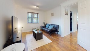 Ein Wohnzimmer mit Flachbildfernseher, schwarzem Ledersofa, Couchtisch und einem Fenster mit Jalousien.