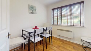 Ein Essbereich mit Tisch, Stühlen, einem roten Untersetzer, einem Gemälde, einem Fenster mit Jalousien und einer Heizung.