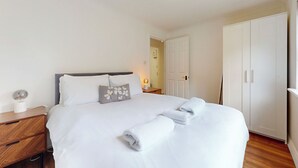 Ein ordentlich bezogenes Bett mit weißen Leinen und einem Zierkissen, ein Nachttisch aus Holz mit einer Lampe und ein weißer Kleiderschrank.