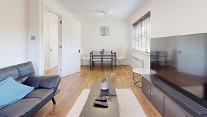 Ein Wohnzimmer mit einer Couch, einem Couchtisch mit Fernbedienungen, einem Esstisch mit Stühlen und einem Fernseher.