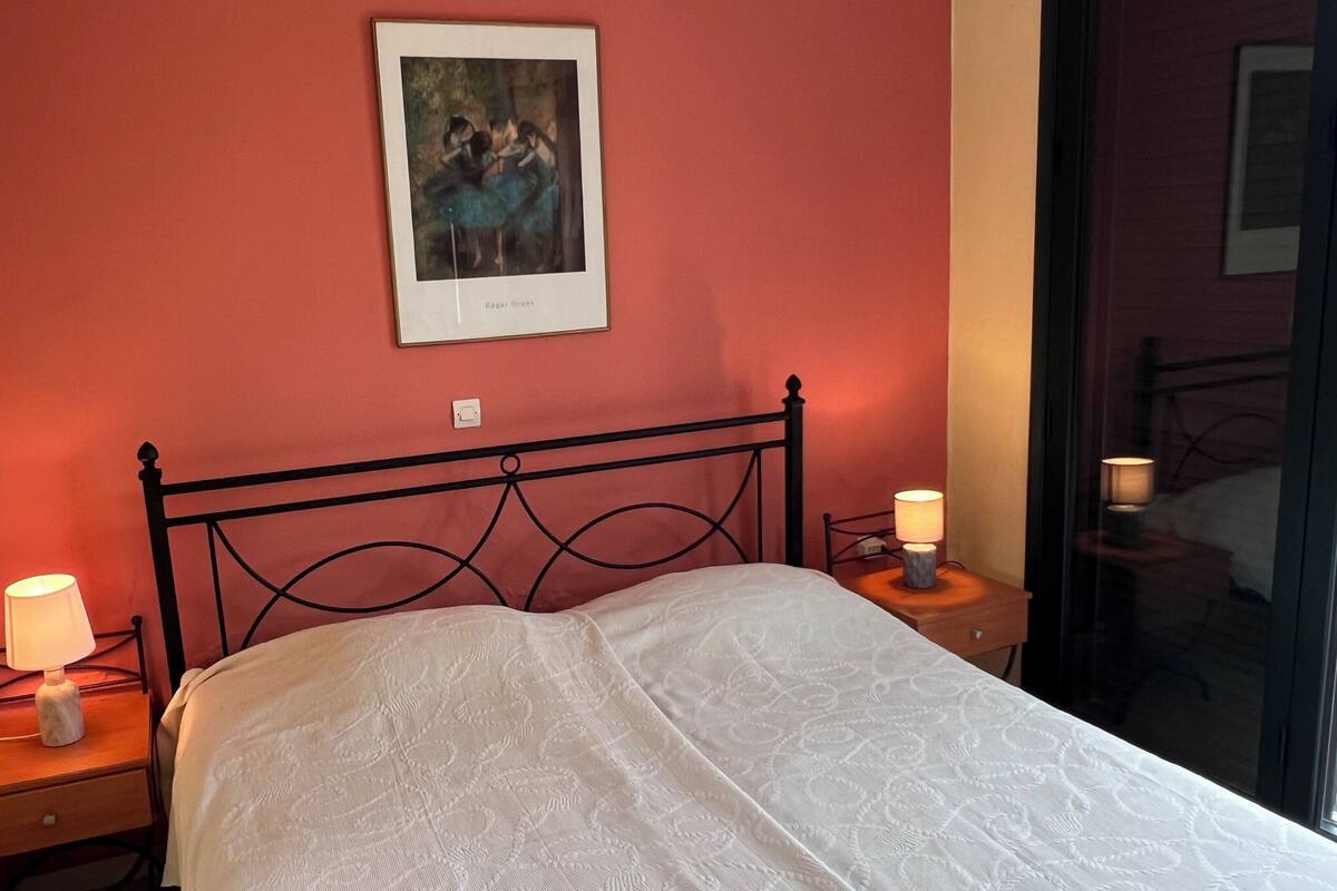 A bed with a white bedspread, two bedside lamps, a framed picture on the wall, and a window with a view.