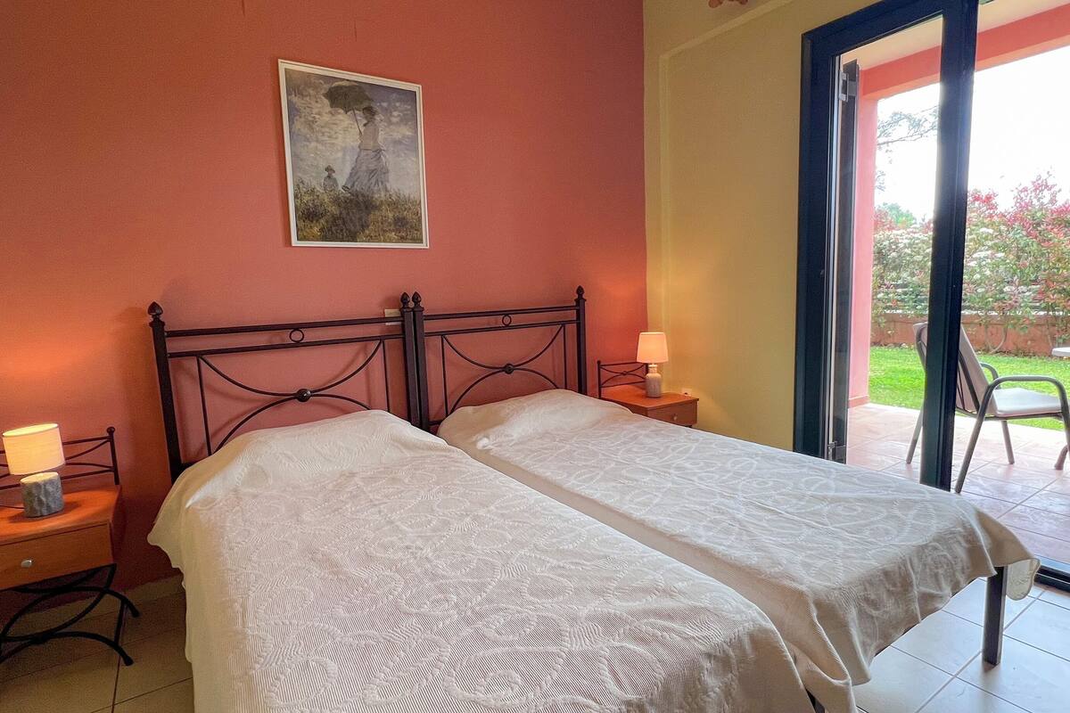 A bedroom with two beds, a nightstand with a lamp, a painting on the wall, and a view of an outdoor patio with chairs and a table.