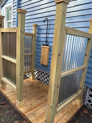 Una terraza de madera con un portón metálico, un poste de madera con una lámpara colgante y una cerca de piquetes blancos.