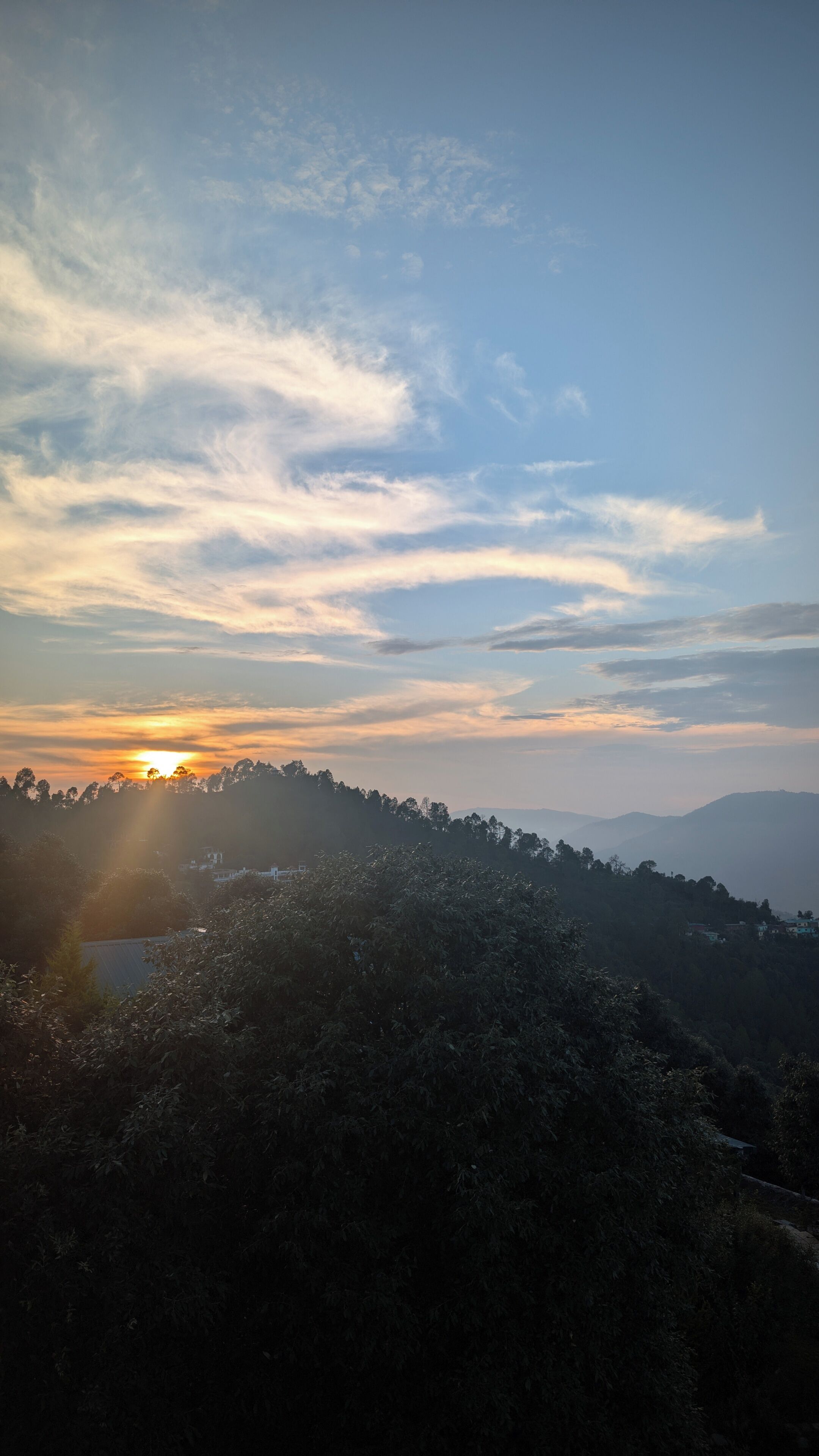 Paesaggio con il sole al tramonto dietro una collina, alberi sullo sfondo e cielo sereno.